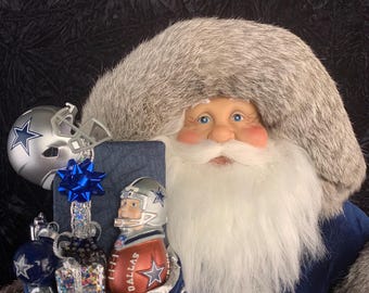Puede incluir: Una figura de Papá Noel con una sudadera gris de los Dallas Cowboys y un abrigo azul. Papá Noel sostiene un saco azul lleno de adornos con temática de fútbol y un balón de fútbol en miniatura. Un casco plateado está encima del saco.