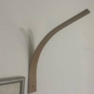 May include: A curved wooden shelf bracket fixed to a white wall. The bracket is a light brown colour and has a rectangular base with two screws. The curved arm extends outwards, providing support for a shelf.