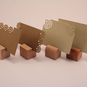 May include: Four decorative place card holders. The cards are in shades of brown and beige, with decorative cut-out edges. They are held by small, wooden blocks. The cards are approximately 10 cm in length.