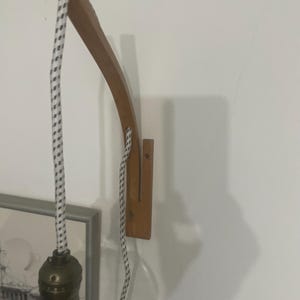 Może przedstawiać: Drewniany kinkiet z zakrzywionym ramieniem. Posiada przezroczystą szklaną żarówkę i czarno-biały sznur w paski. Drewniane ramię jest przymocowane do prostokątnej drewnianej podstawy. Kinkiet jest zamontowany na białej ścianie.