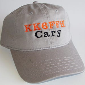 May include: A gray baseball cap with a curved brim. The cap features embroidered text in orange and black: "KK6FFH Cary". The hat is made of a soft fabric and has a button on top.