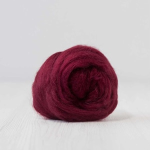 May include: A ball of deep burgundy wool roving, tightly wound and textured. The wool is a rich, saturated color, and the fibers are visible. The background is a soft white, providing a contrast to the dark wool.