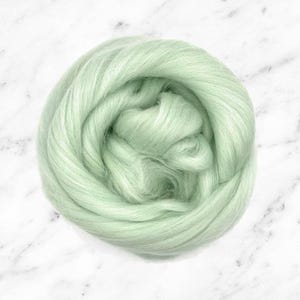 May include: A circular bundle of pale green wool roving, set against a white marble surface. The wool appears soft and fluffy, with a subtle sheen. This material is suitable for various fibre art projects, such as needle felting and spinning, and is measured in grams.