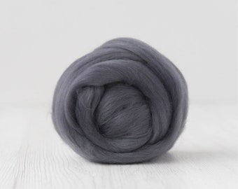Op de afbeelding: Een bal donkergrijze wolvlies, strak opgerold en gecentreerd tegen een witte, getextureerde achtergrond. De wol lijkt zacht en donzig, ideaal voor diverse vezelkunstprojecten zoals vilten of spinnen.