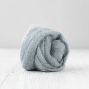 May include: A ball of pale blue roving yarn, tightly wound and textured. The yarn has a soft, muted hue, and appears fluffy. The background is plain white, highlighting the yarn's colour and texture. Suitable for various craft projects.