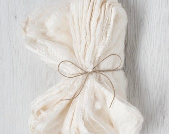 May include: A folded, off-white, semi-sheer fabric runner with a crinkled, textured surface. The fabric is neatly bundled and secured in the center with a thin, natural-colored jute twine tied into a simple bow, creating a gathered appearance.