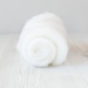 May include: A rolled bundle of soft, white material, possibly wool or cotton, rests on a light wooden surface. The material has a fluffy texture and a spiral pattern is visible. The background is a plain, light grey.