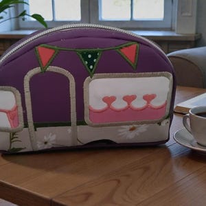 May include: A purple campervan-shaped travel bag with a zip closure. The bag has a decorative design of a camper with a window, hearts, and daisy accents. A cup of coffee sits on a saucer on a wooden table.