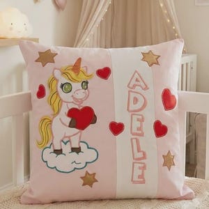 Peut inclure: Coussin carré rose avec un motif de licorne. La licorne a une crinière et une corne jaunes, tenant un cœur rouge. Le coussin comporte également le nom "ADELE" en lettres roses, des cœurs rouges et des étoiles dorées.