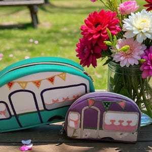 Peut inclure: Deux trousses de maquillage zippées en forme de caravanes vintage. L'une est turquoise avec un motif de fanions blancs et oranges, l'autre est violette avec des fanions roses et verts. Un bouquet de fleurs est dans un bocal en verre.