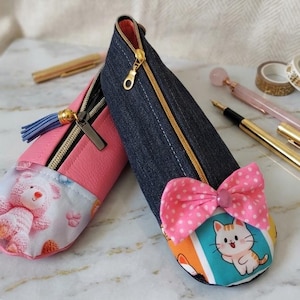 Op de afbeelding: Twee etuis met rits. De ene is roze met een teddybeerprint en een blauw kwastje. De andere is van donkerblauwe denim met een roze strik met stippen en een cartoon kattenprint. Beide etuis zijn voorzien van goudkleurige ritsen.