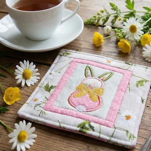 May include: A square, quilted coaster with a pink bunny design and a yellow bow. The coaster is surrounded by a pink border with white polka dots and a floral pattern. A white teacup and saucer are in the background, along with daisies and yellow flowers.