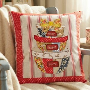 Peut inclure: Un coussin carré avec une applique centrale représentant trois bols à mélanger empilés surmontés d'un nœud floral jaune. Les bols sont décorés de motifs rouges, blancs et bleus avec des étiquettes rouges portant les mots Home, Sweet, Home.