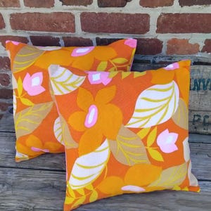 Op de afbeelding: Twee vierkante kussens met een levendige oranje achtergrond en een bloemenpatroon. Het ontwerp heeft grote oranje bloemen, witte en bruine bladeren en roze accenten. De kussens liggen op een houten oppervlak.