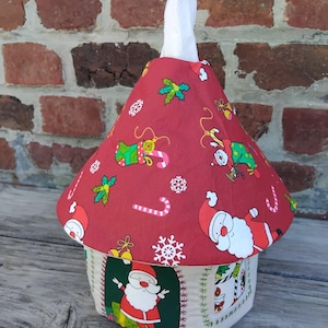 Könnte beinhalten: Eine Stoffhülle für Taschentuchboxen in Hausform mit einem konischen roten Dach, das mit einem Weihnachtsmuster aus Weihnachtsmann, Zuckerstangen und Schneeflocken bedruckt ist. Die Basis ist cremefarben mit grünen Feldern und Weihnachtsmannmotiven.
