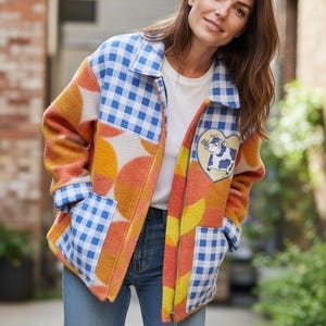 Puede incluir: Chaqueta patchwork colorida con cuello y detalles de bolsillo de cuadros azules. La chaqueta presenta una mezcla de patrones geométricos naranjas, amarillos y blancos, y un parche en forma de corazón con una ilustración de vaca. El modelo lleva vaqueros azules.