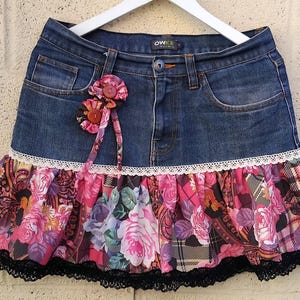 Peut inclure: Une jupe en jean avec un volant patchwork floral. La jupe présente un denim délavé foncé avec une bordure en dentelle blanche et un ourlet en dentelle noire. Une fleur en tissu est attachée à l'avant.