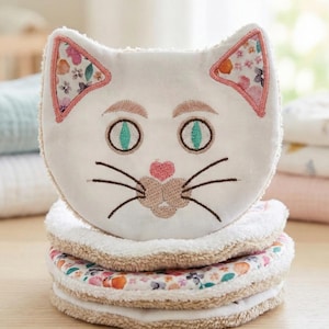 Peut inclure: Une pile de débarbouillettes blanches et à motifs floraux, surmontée d'un visage de chat. Le visage du chat présente des détails brodés, notamment des yeux turquoise, des moustaches et des motifs floraux sur les oreilles. Les débarbouillettes sont en matière douce et absorbante.