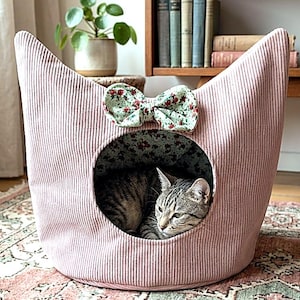 May include: A soft pink corduroy cat bed featuring a circular entrance and a decorative bow with a green and red floral pattern. A grey tabby cat is resting inside the bed, which has a matching floral interior lining and pointed top edges.