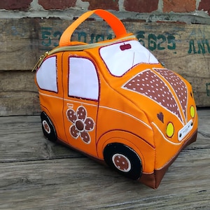 May include: Orange car-shaped lunch bag with a brown flower and star pattern. The bag has white window accents, black wheels, and an orange handle. The zipper and trim are also orange. The bag is sitting on a wooden surface.