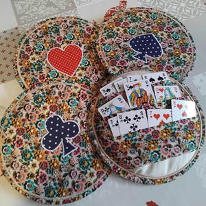 May include: A set of four circular fabric card holders featuring a colorful floral pattern. Each holder displays a different white-dotted suit symbol: heart, spade, club, and diamond. One holder is open, revealing a deck of playing cards inside.