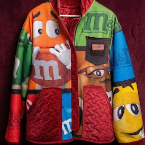 Könnte beinhalten: Eine farbenfrohe Patchwork-Jacke mit M&M's-Charakteren und -Marken. Die Jacke hat ein rotes, gestepptes Futter und Taschen, mit einem Lee-Etikett auf der Brusttasche. Das Design integriert orange, grüne, blaue und gelbe Paneele.