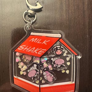 May include: A clear acrylic keychain shaped like a milk carton, featuring the words "MILK SHAKE" in red. Inside are cow-shaped charms and heart-shaped confetti. The keychain has a silver clasp.