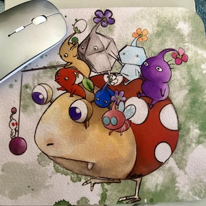 May include: A colorful mousepad featuring a cartoon illustration of a large, yellow, round creature with a red mushroom cap. The creature is surrounded by smaller, colorful creatures with various shapes and designs.