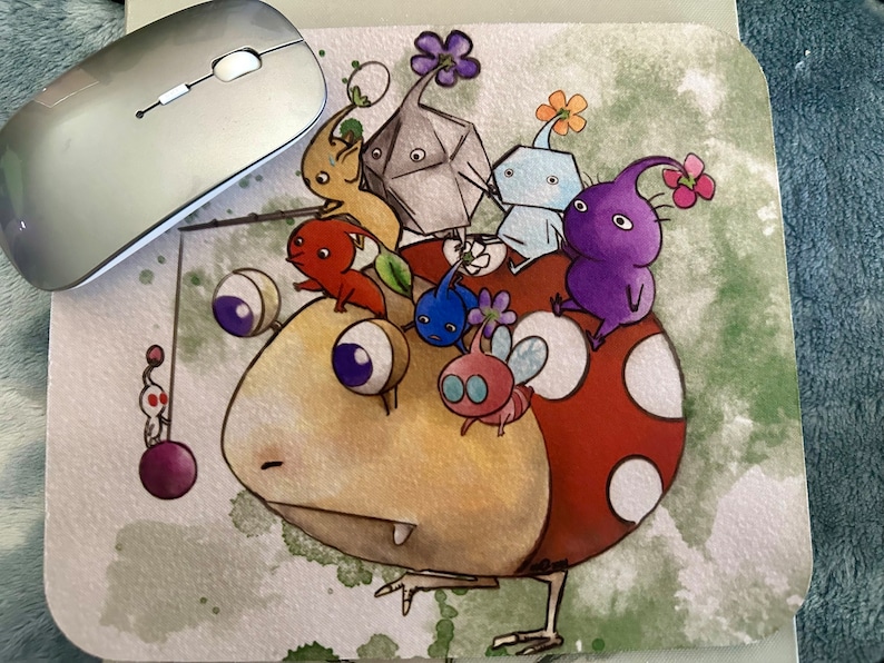 May include: A colorful mousepad featuring a cartoon illustration of a large, yellow, round creature with a red mushroom cap. The creature is surrounded by smaller, colorful creatures with various shapes and designs.