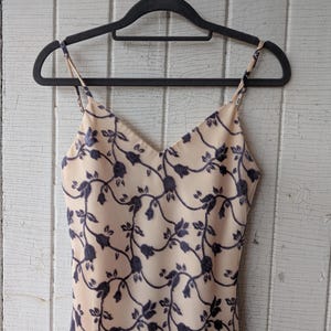 May include: A beige slip dress with a dark blue floral pattern, hanging on a black hanger. The dress has thin straps and a V-neckline. The floral design features winding vines and leaves.