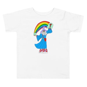 Może przedstawiać: Biały t-shirt z kolorową ilustracją czarodzieja trzymającego różę i zieloną kulę. Czarodziej stoi na platformie z tęczą łukiem nad nim. Tekst "Love Billy Light" jest napisany pod platformą.