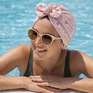 May include: A light pink turban with a bow detail and floral pattern. The person is wearing cream-colored sunglasses with brown lenses and a green swimsuit. The background is a blue swimming pool.