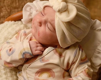 May include: A baby wearing a white crocheted hat with a bow on top and a white onesie with a rainbow pattern.