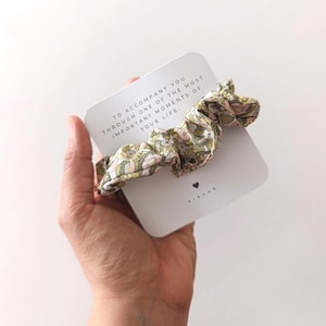 May include: A floral scrunchie in shades of green, pink, and yellow rests on a white card. The card has text that reads, "TO ACCOMPANY YOU THROUGH ONE OF THE MOST IMPORTANT MOMENTS OF YOUR LIFE." A small black heart is visible.