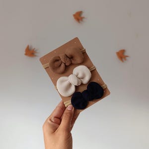 Peut inclure: Ensemble de trois bandeaux pour bébé avec des nœuds décoratifs de couleur beige, crème et bleu marine. Les nœuds sont fixés à de fines bandes élastiques neutres et présentés sur une carte marron. Fond beige clair avec des décorations en forme d'oiseaux orange.