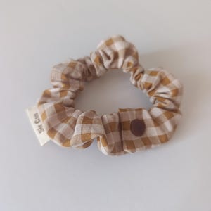 May include: A tan and white checkered scrunchie with a brown button closure. The scrunchie has a small white tag that reads "Sir Cus". This hair accessory is a stylish addition to any hairstyle.