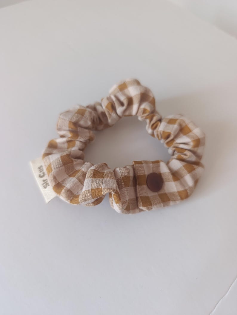 May include: A tan and white checkered scrunchie with a brown button closure. The scrunchie has a small white tag that reads "Sir Cus". This hair accessory is a stylish addition to any hairstyle.
