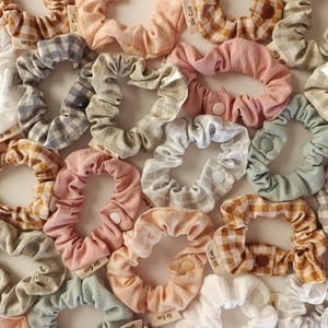May include: A collection of assorted scrunchies in various colors and patterns, including gingham and solid fabrics. The scrunchies are in shades of white, pink, gray, and peach. Each scrunchie has a small tag attached.