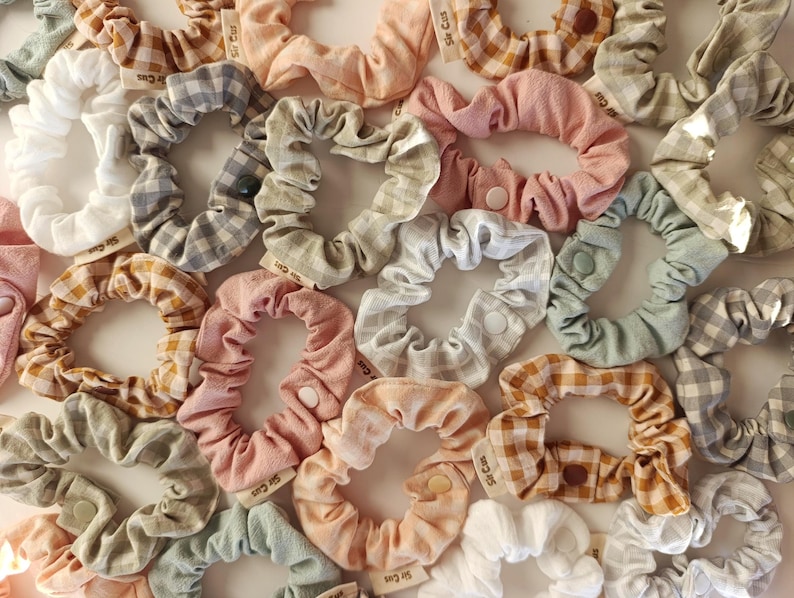 May include: A collection of assorted scrunchies in various colors and patterns, including gingham and solid fabrics. The scrunchies are in shades of white, pink, gray, and peach. Each scrunchie has a small tag attached.
