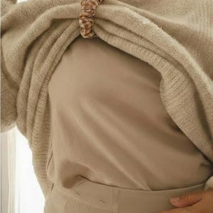 May include: A beige sweater is held together by a brown and white checkered scrunchie. The sweater is draped over a beige top and pants. The outfit is a neutral color palette.