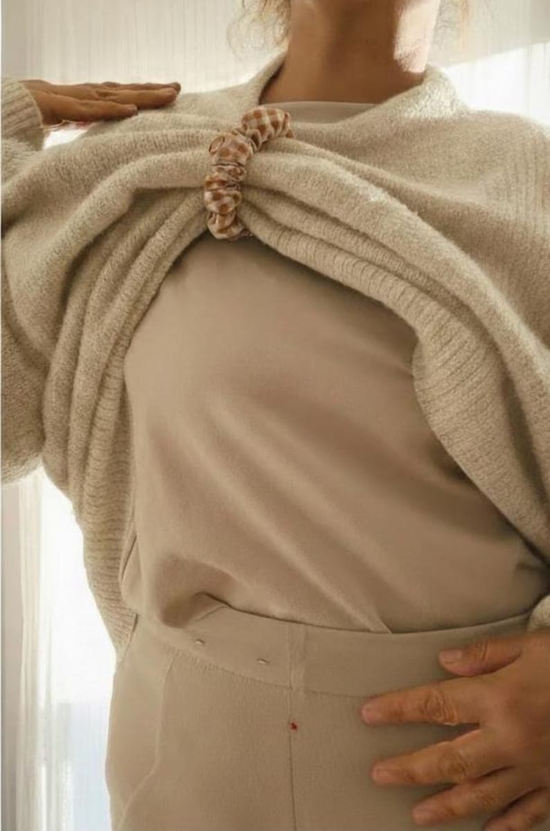 May include: A beige sweater is held together by a brown and white checkered scrunchie. The sweater is draped over a beige top and pants. The outfit is a neutral color palette.