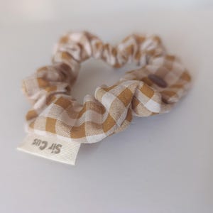 May include: A scrunchie made of fabric with a tan and white gingham pattern. A small white tag with the text "Sir Cus" is attached. The scrunchie is a hair accessory.
