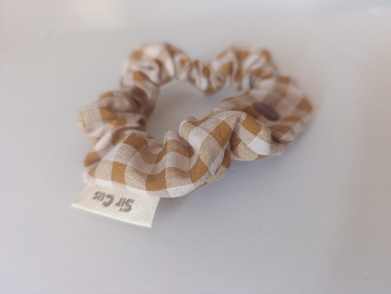 May include: A scrunchie made of fabric with a tan and white gingham pattern. A small white tag with the text "Sir Cus" is attached. The scrunchie is a hair accessory.