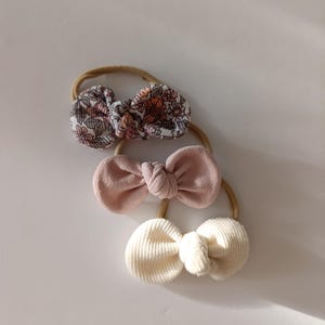 Könnte beinhalten: Drei Baby-Haarbänder mit Schleifenakzenten. Eine Schleife hat ein Blumenmuster in Rosa, Orange und Grau. Die zweite Schleife ist einfarbig rosa, und die dritte ist cremefarben. Jede Schleife ist an einem neutralen Gummiband befestigt.