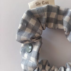 May include: A gray and white gingham scrunchie with a gray button closure. The scrunchie has a small tag that reads "Sir Cus". The fabric is a soft cotton material.