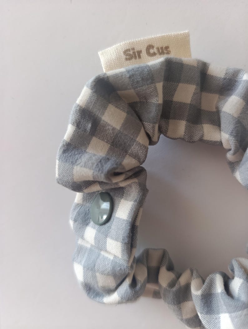 May include: A gray and white gingham scrunchie with a gray button closure. The scrunchie has a small tag that reads "Sir Cus". The fabric is a soft cotton material.