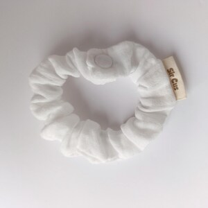 Puede incluir: Un coletero de tela blanca con forma circular y material fruncido. Una peque&ntilde;a etiqueta de tela beige est&aacute; unida al lateral con el texto Sir Cus impreso en marr&oacute;n. Un solo bot&oacute;n de presi&oacute;n blanco es visible en la parte superior.
