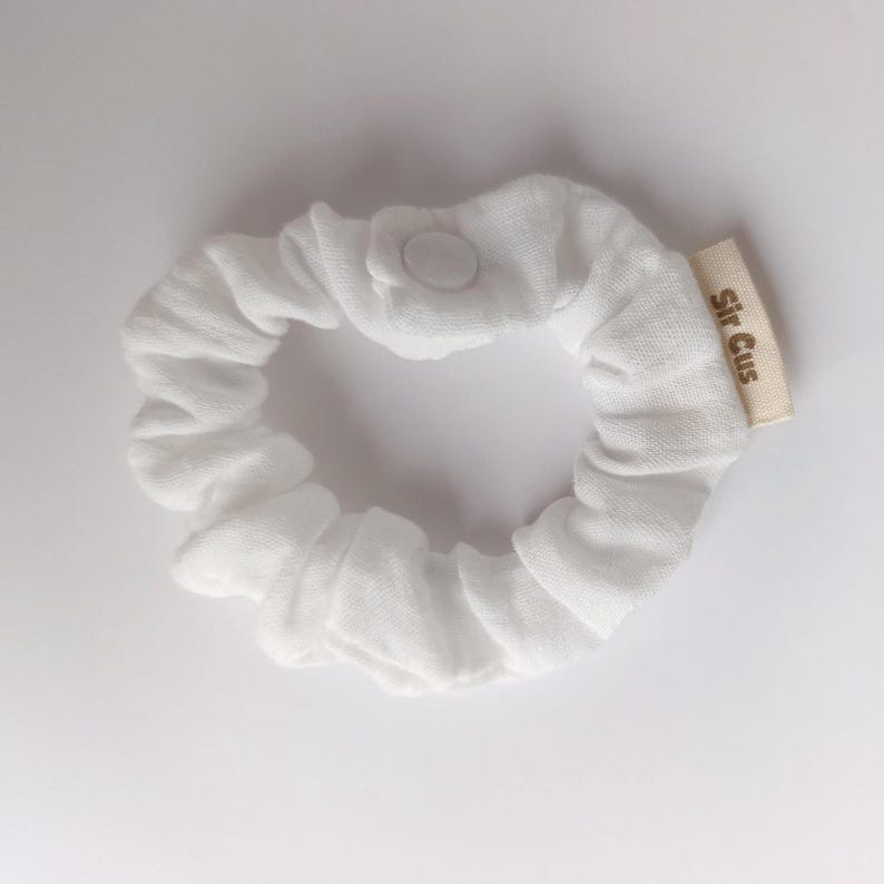 Puede incluir: Un coletero de tela blanca con forma circular y material fruncido. Una peque&ntilde;a etiqueta de tela beige est&aacute; unida al lateral con el texto Sir Cus impreso en marr&oacute;n. Un solo bot&oacute;n de presi&oacute;n blanco es visible en la parte superior.