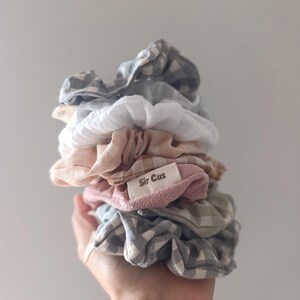 May include: A stack of assorted scrunchies in various colors and patterns, including gingham and solid hues. The scrunchies are in shades of white, pink, gray, and blue. A small tag with the text "Sir Cus" is visible.