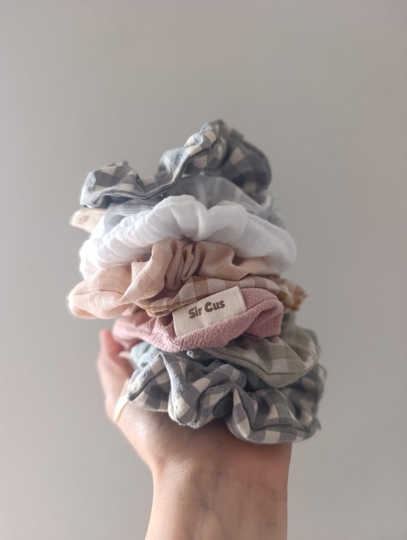 May include: A stack of assorted scrunchies in various colors and patterns, including gingham and solid hues. The scrunchies are in shades of white, pink, gray, and blue. A small tag with the text "Sir Cus" is visible.